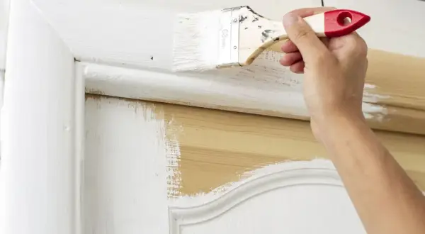 Main d'une personne peignant une porte en bois avec un pinceau. La peinture blanche recouvre partiellement le bois naturel. Quel rouleau pour peindre une porte en bois ?