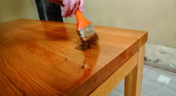 Application d'une huile dure sur un plateau en bois. Une main tient un pinceau orange pour appliquer le produit, révélant la beauté naturelle du bois. Recette huile dure.