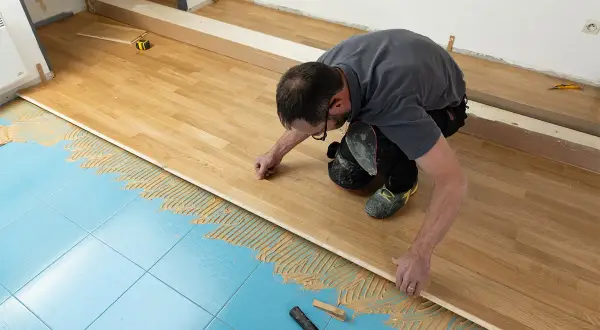 Un homme pose du parquet massif sur un sol bleu carrelé, appliquant de la colle. Il est en train de réaliser un projet de rénovation.