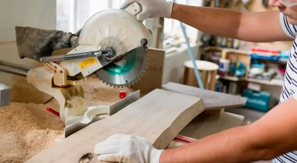 Un artisan utilise une scie à onglet pour couper une planche de bois. Il tient la pièce avec des gants, prêt à réaliser une coupe précise.