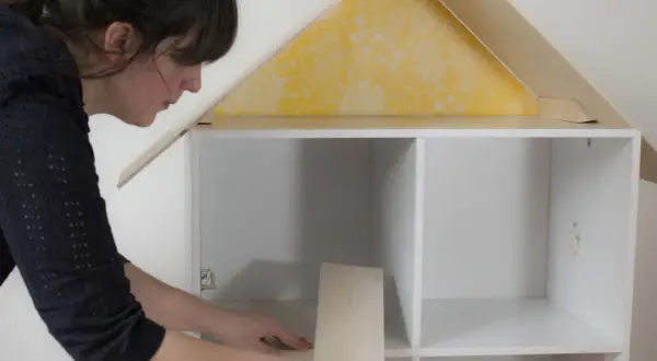Une femme assemble une maison de poupée. Elle pose une pièce en bois dans une étagère blanche, sous un toit jaune à motifs.