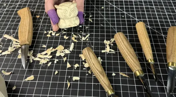 Une main tient une petite chouette en bois sculpté, entourée d'outils et de copeaux. Idéal pour une **débutant sculpture bois facile**.