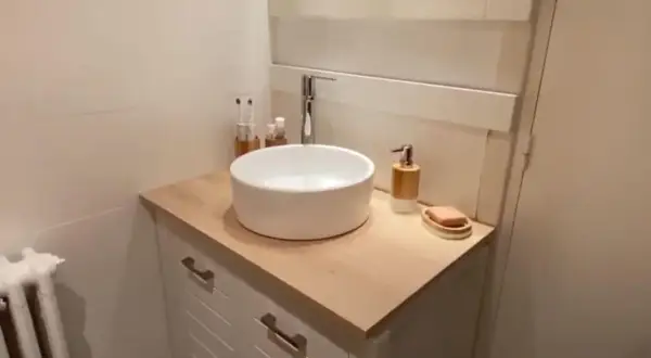 Un meuble de salle de bain relooké avec un plan de travail en bois clair, une vasque blanche et des accessoires modernes.
