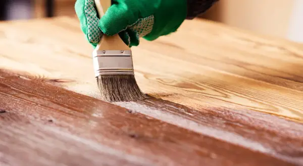Application d'une couche de vernis sur un plateau en bois. La main gantée de vert applique le produit avec un pinceau, révélant la beauté naturelle du bois.