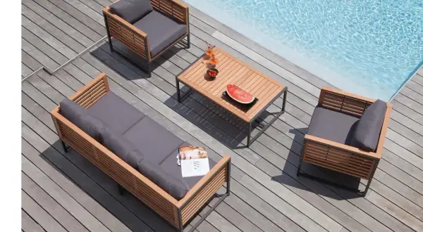 Salon de jardin en bois teck avec coussins gris, près d'une piscine. Table basse avec fruits et boissons.