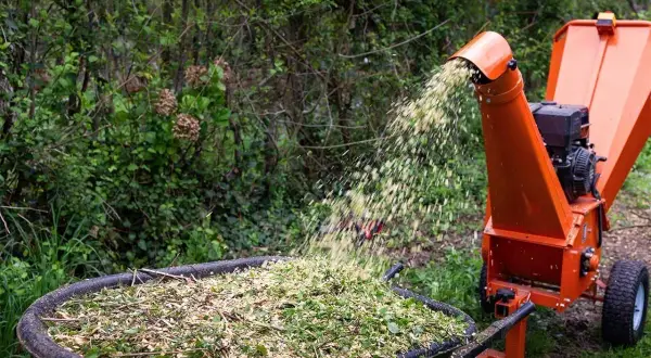 Une machine à faire des copeaux de bois orange projette des débris végétaux dans une brouette remplie de copeaux.