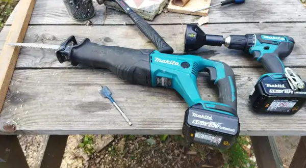 Outils de bricolage sur une table en bois : scie sabre et perceuse de marque makita, marteau, clous, mèche.