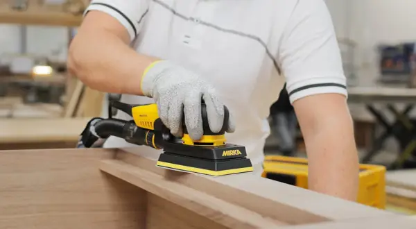 Main gantée utilise une ponceuse Mirka pour poncer une porte en bois, préparant la surface pour une finition parfaite.