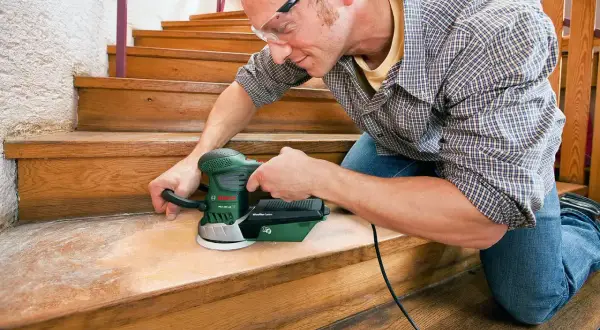 Un homme utilise une ponceuse pour rénover les marches d'un escalier en bois.