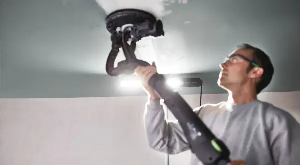 Un homme utilise une ponceuse de plafond Festool, éclairée par une lampe rasante Festool, pour lisser une surface.