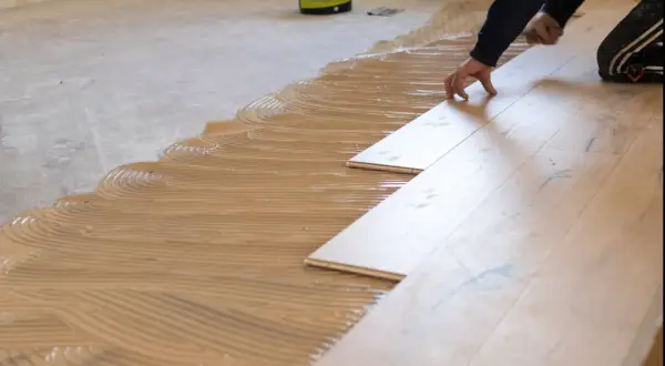 Pose de parquet collé : application de colle en plein à la spatule crantée, avant la pose des lames de bois.
