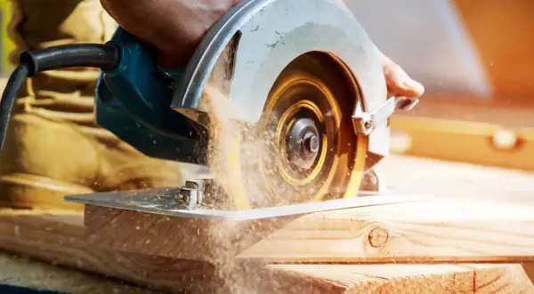 Main d'un artisan guidant une scie circulaire. Le sens de rotation de la lame de scie circulaire projette de la sciure.