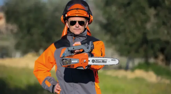 Un homme en tenue de protection, casque et lunettes, présente une tronçonneuse thermique. Il vous aide à choisir la meilleure.