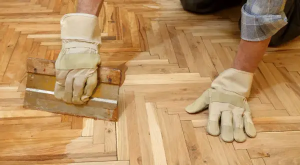Un homme, ganté, applique de la cire sur un parquet en chevrons avec une raclette.