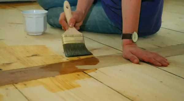 Application d'une couche protectrice sur un parquet huilé ou ciré. Une main tient un pinceau, l'autre est posée sur le bois.