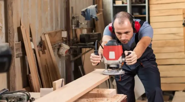 Artisan concentré sur ses réalisations à la défonceuse, créant des formes précises sur une planche de bois dans son atelier.