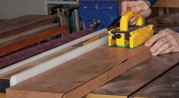 Maintenant, je vais fabriquer une scie sur table. Un artisan utilise un poussoir jaune pour guider une planche de bois sur la scie.