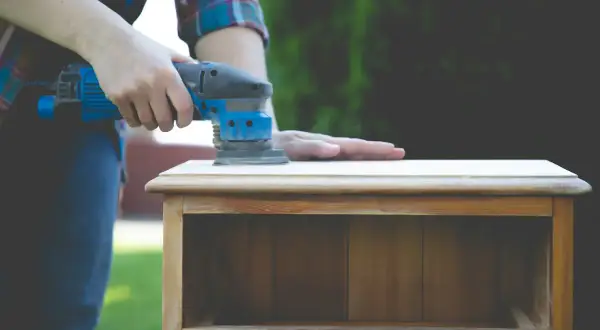 Une personne utilise une ponceuse pour lisser le dessus d'une petite commode en bois, un travail qui demande de poncer endroit difficile.