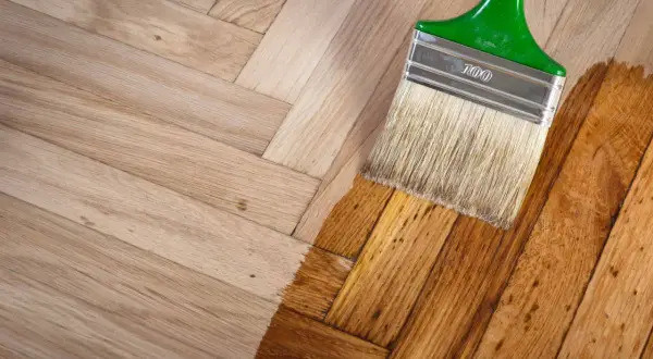 Un pinceau applique une couche de vernis sur un parquet en bois clair, transformant sa teinte. Le processus de nettoyer parquet est en cours.