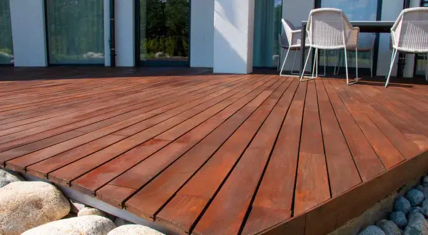 Belle terrasse bois sur plot béton, avec des galets et des gros rochers. Des chaises et une table sont visibles au fond.