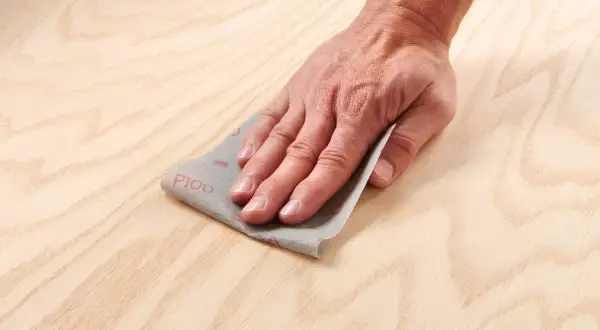 Main d'artisan effectuant un égrenage sur une surface en bois clair avec du papier de verre P100.