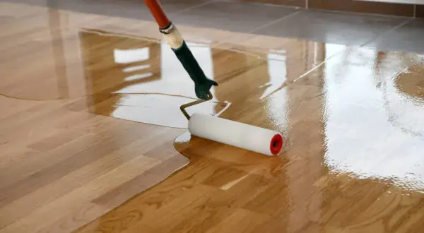 Application d'une première couche de vitrificateur sur un parquet en bois. Le rouleau laisse une surface brillante et humide.