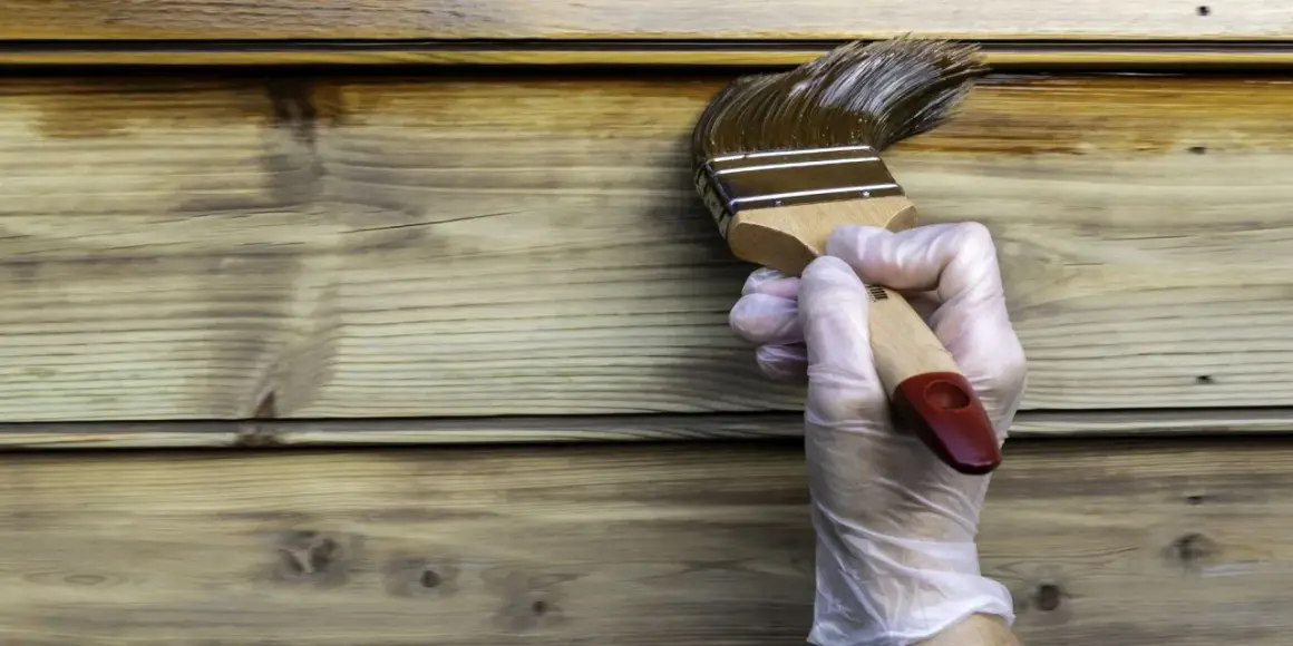 Maintenant, peut on mettre de la lasure sur de la peinture ? Un pinceau applique une lasure foncée sur un bardage en bois clair.