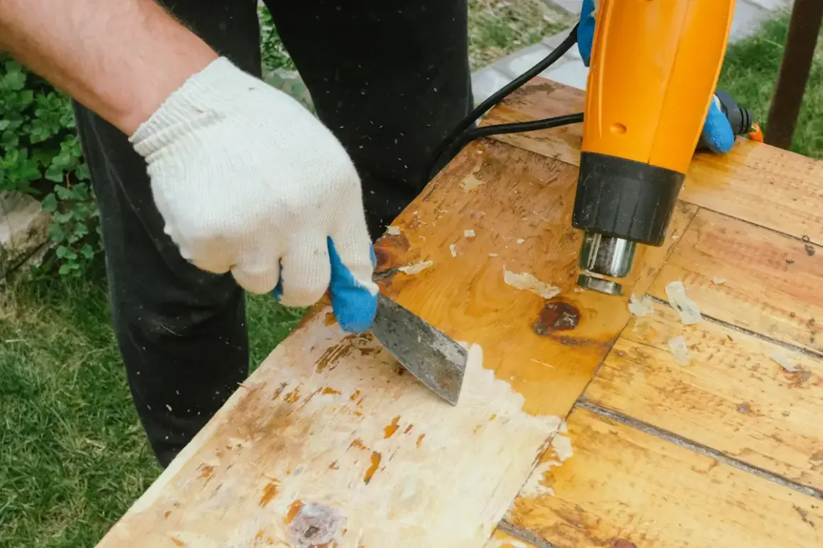 Ganté, une personne utilise un décapeur thermique et un grattoir pour enlever la colle sur du bois.
