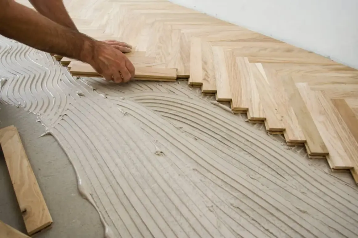 Pose d'un parquet en chevrons sur un lit de colle. Les lames de bois clair sont posées avec soin, formant un motif élégant.