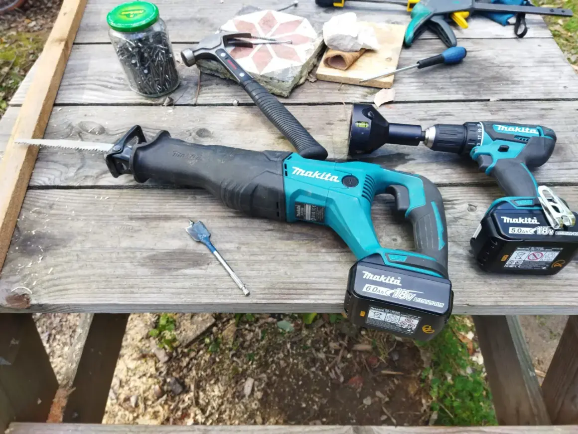 Outils de bricolage sur une table en bois : scie sabre et perceuse de marque makita, marteau, clous, mèche.