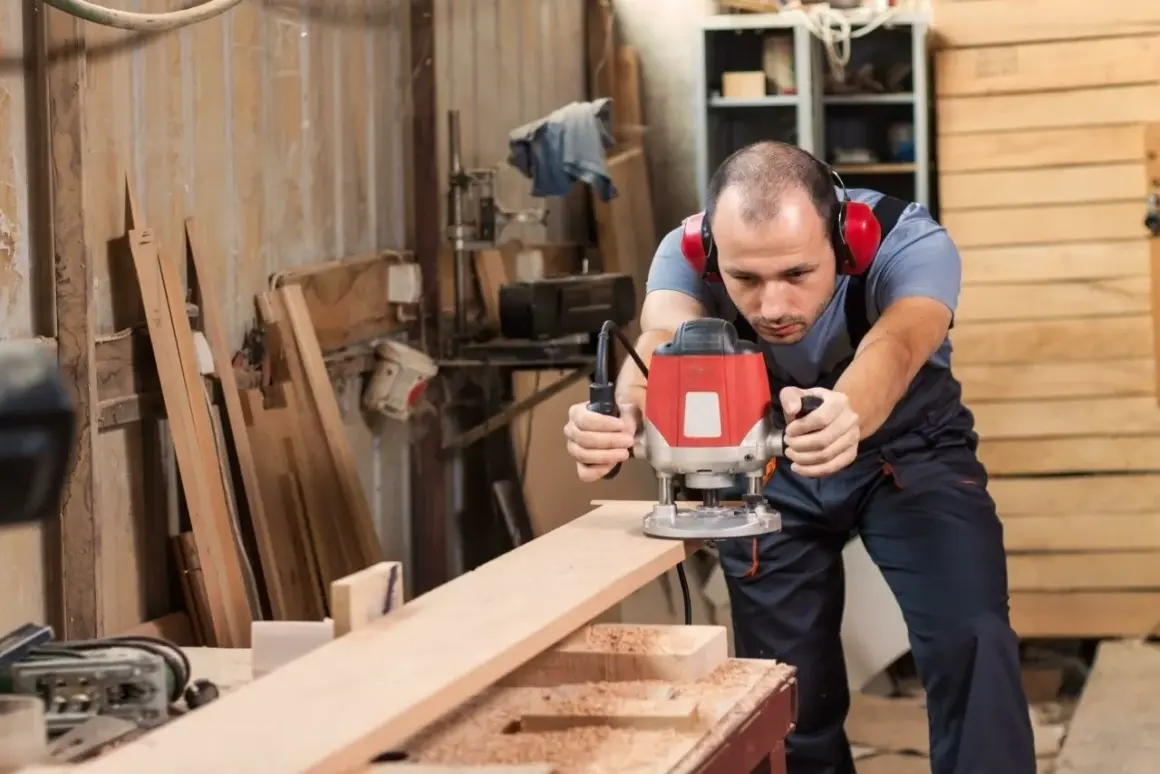 Artisan concentré sur ses réalisations à la défonceuse, créant des formes précises sur une planche de bois dans son atelier.