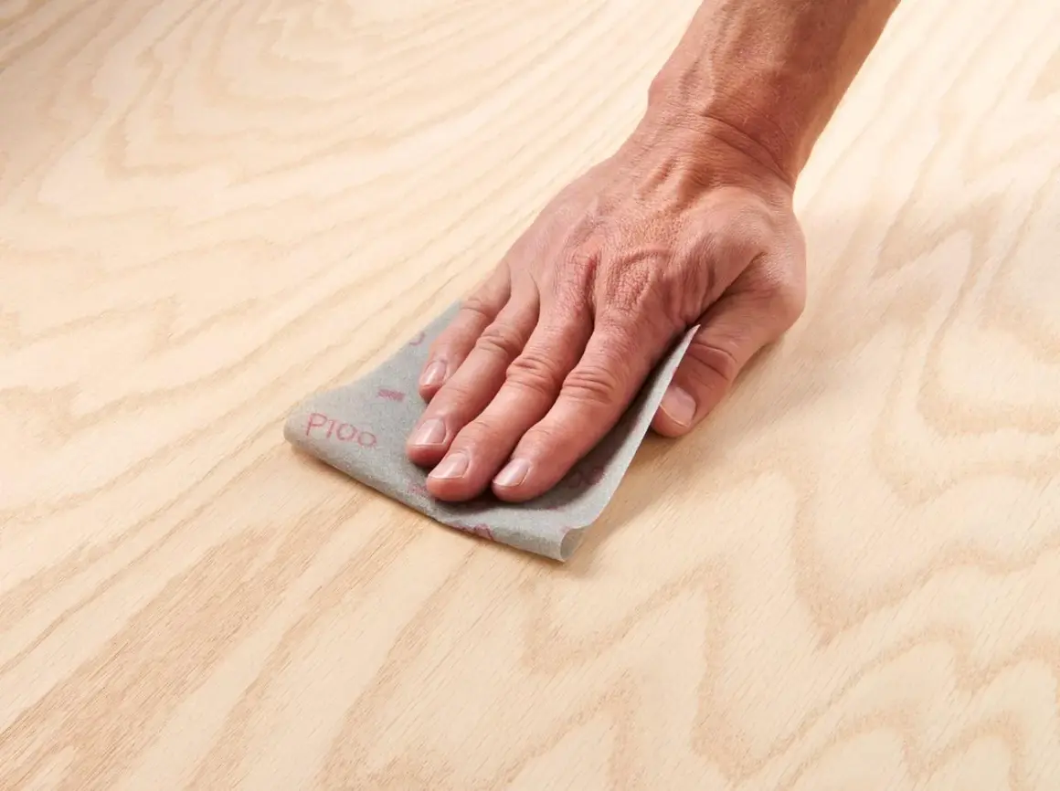 Main d'artisan effectuant un égrenage sur une surface en bois clair avec du papier de verre P100.