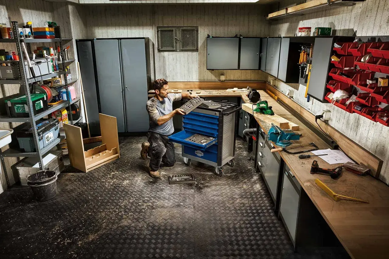 Un homme organise son **aménagement atelier bricolage**. Des outils, des armoires et un chariot à outils remplissent l'espace.