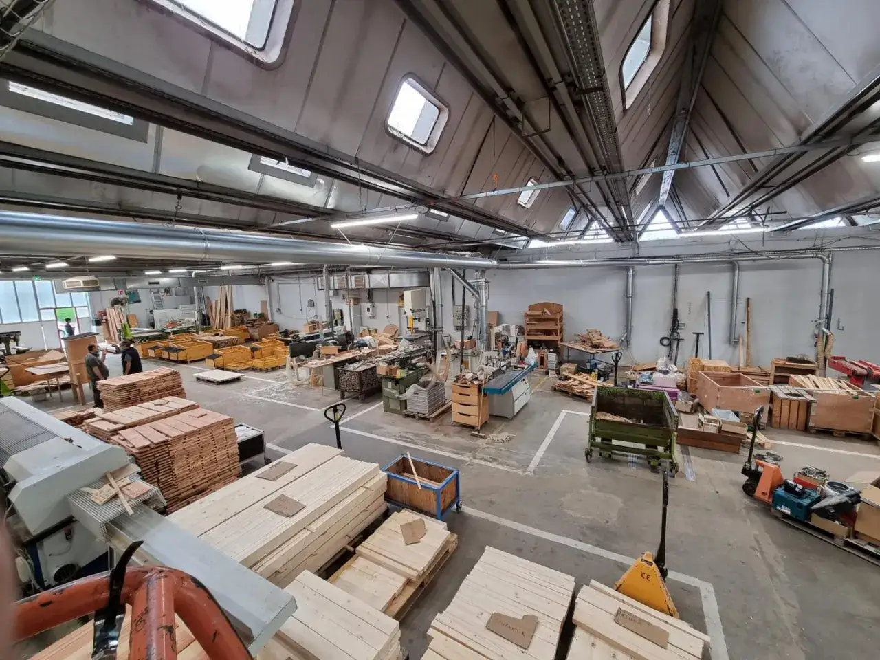 Un grand atelier de menuiserie où l'on peut faire un atelier. Des piles de bois, des machines et des palettes sont visibles.