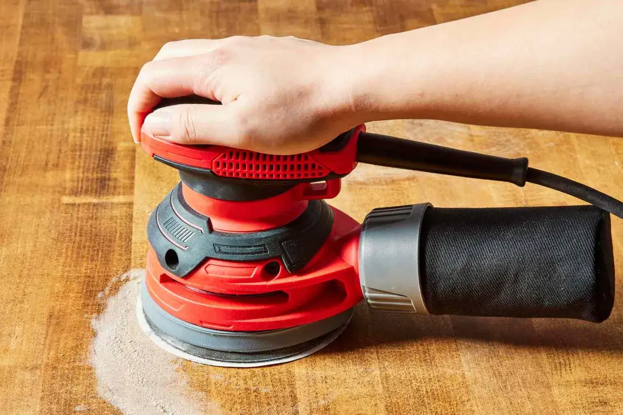 Main d'une personne utilisant une ponceuse de rénovation rouge sur une surface en bois, créant de la poussière.