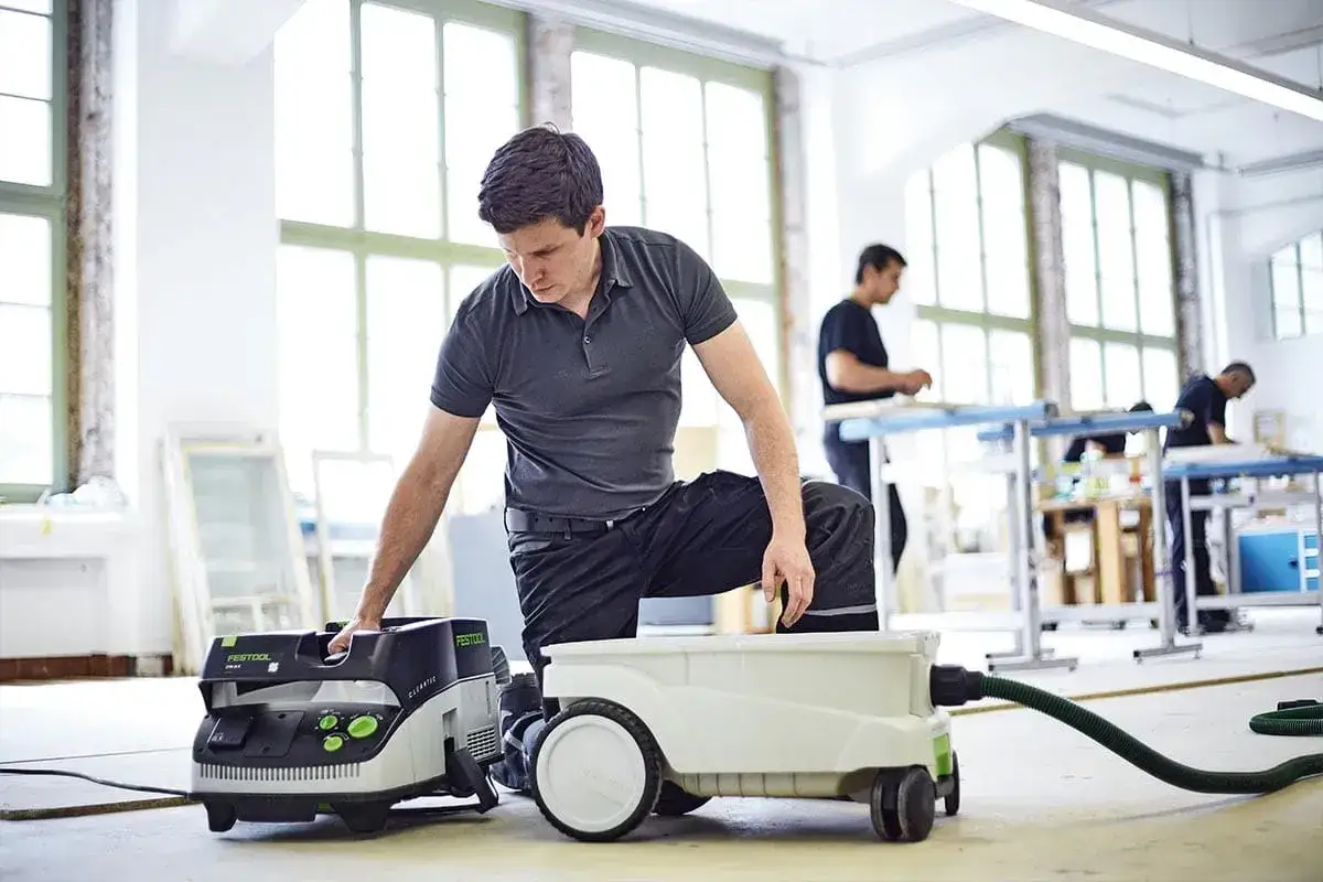 Un homme ajuste son aspirateur Festool CTL 36 E AC dans un atelier lumineux. D'autres travaillent en arrière-plan.