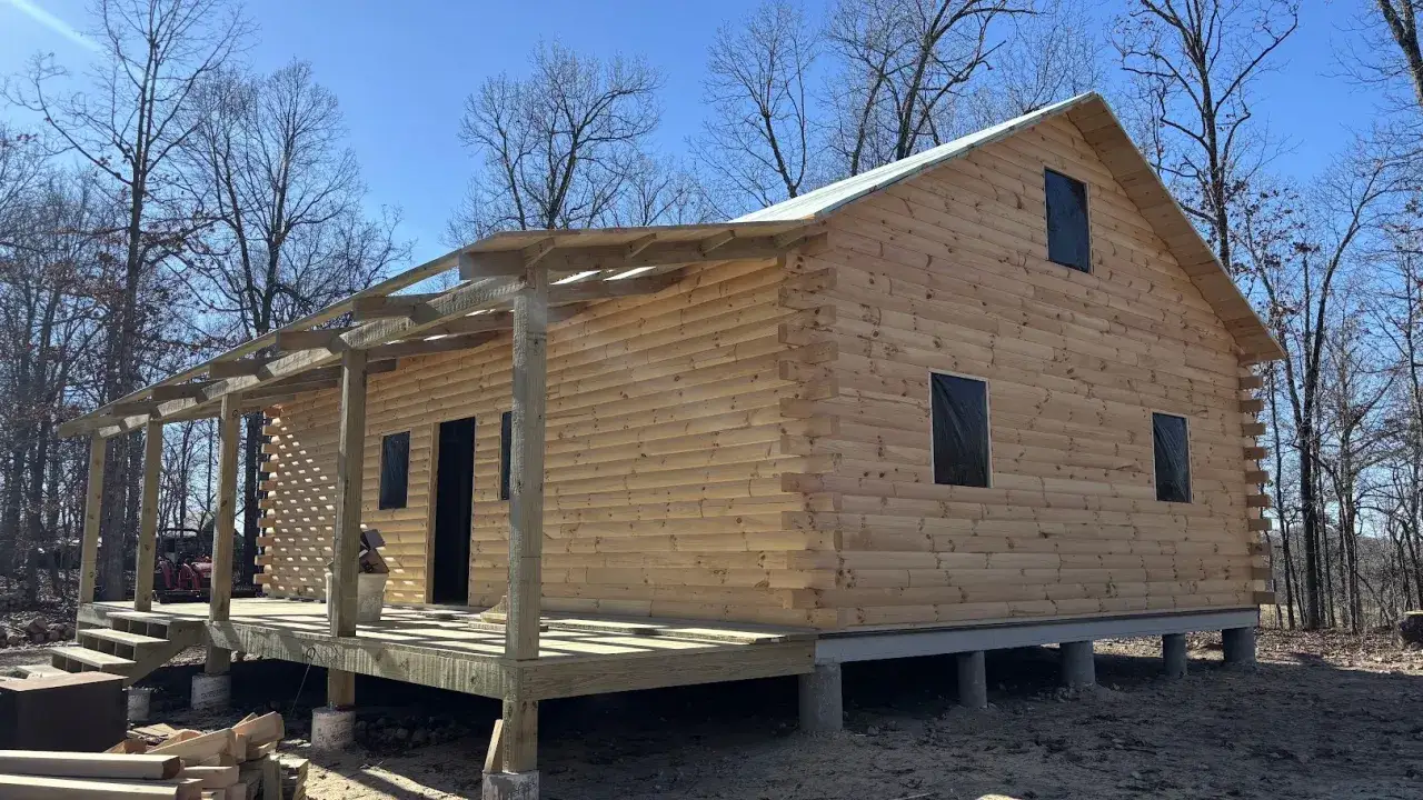 Une cabane en bois fait maison, en construction, avec une grande v&eacute;randa et des fen&ecirc;tres b&acirc;ch&eacute;es, entour&eacute;e d'arbres d&eacute;nud&eacute;s sous un ciel bleu.