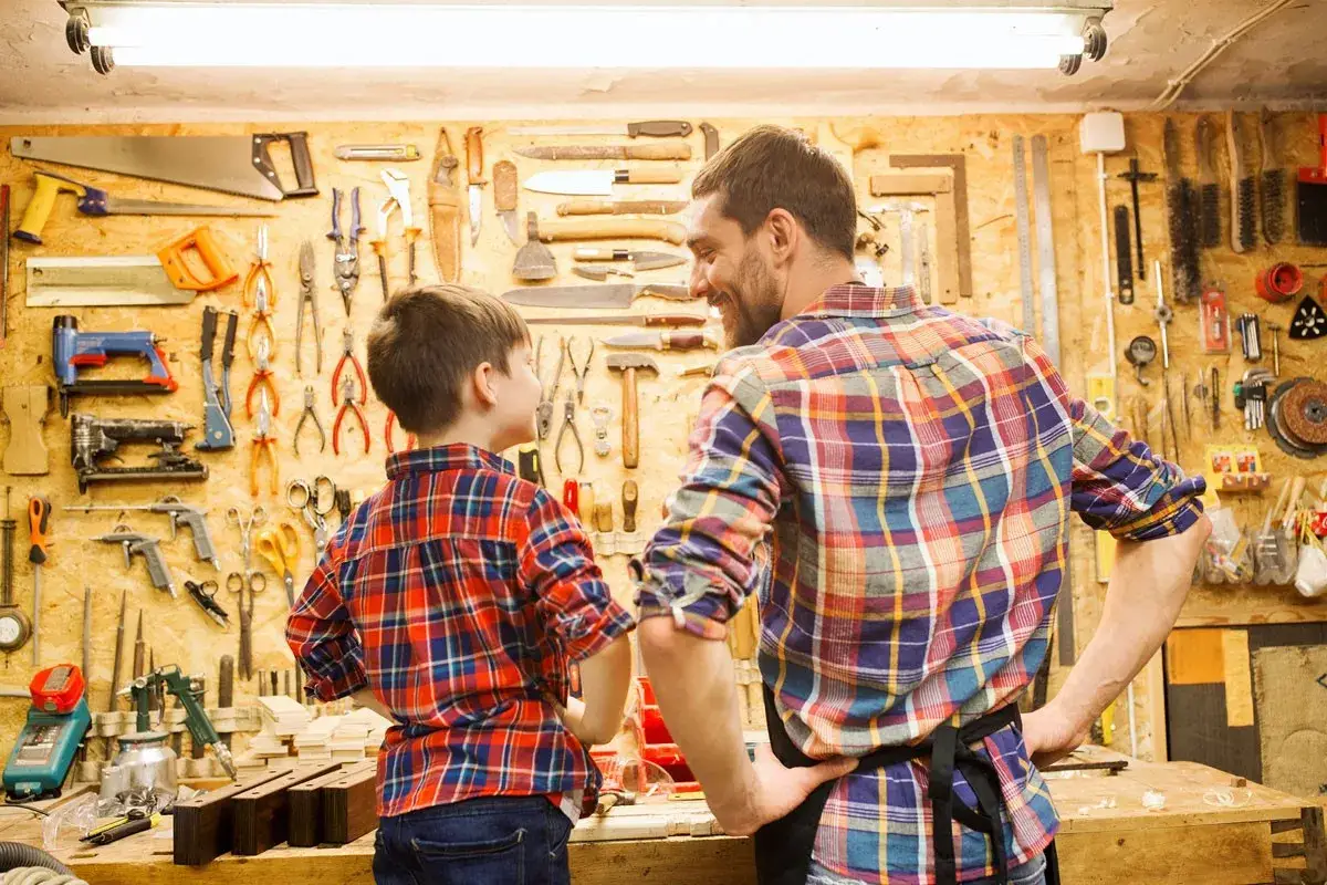Père et fils dans un atelier bricolage, entourés d'outils. Le garçon regarde son père, prêt à apprendre.