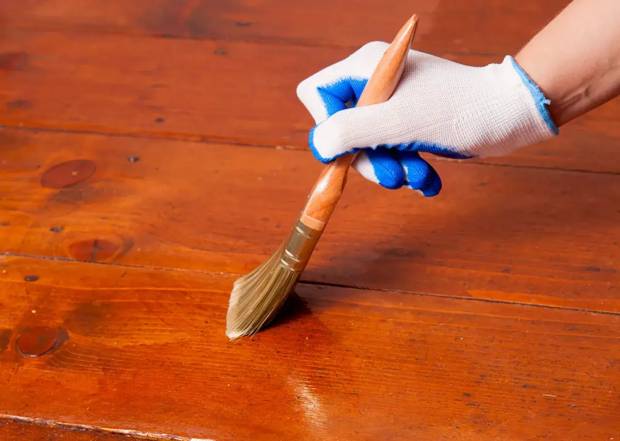 Main d'une personne portant un gant blanc et bleu, appliquant de l'huile sur un parquet en bois.