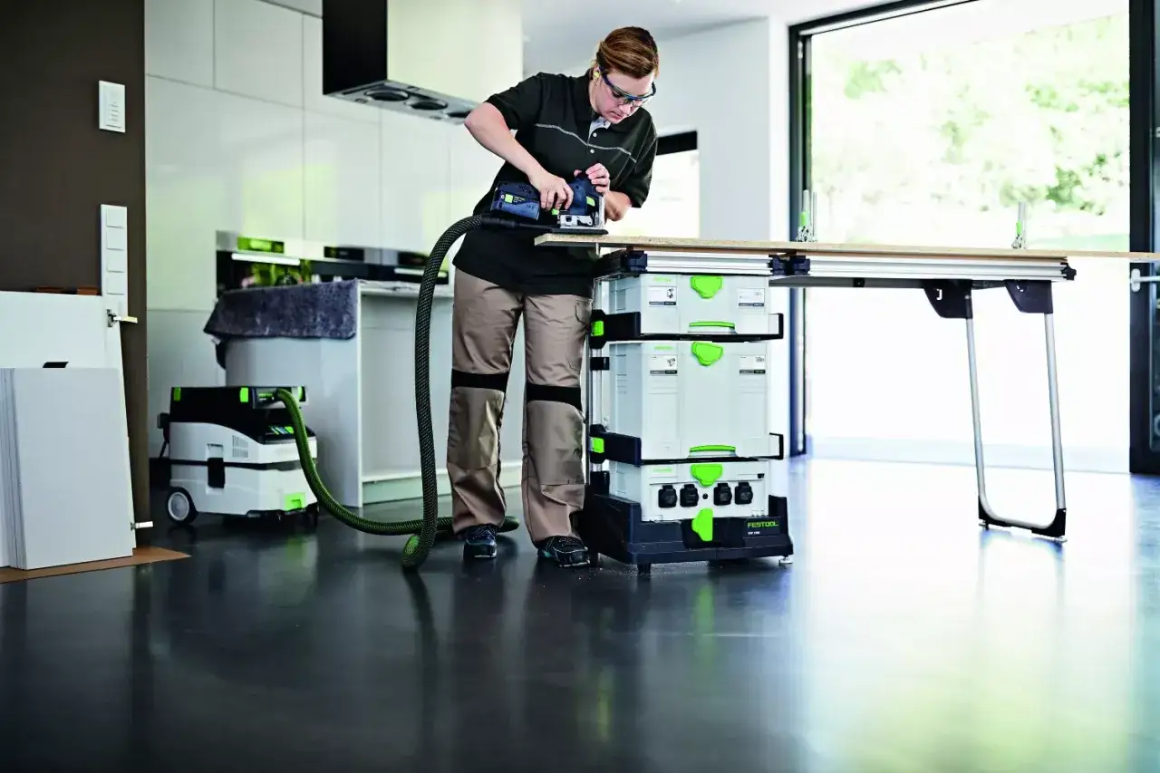Une femme utilise un ponceuse sur un établi, avec un aspirateur et des boîtes de rangement Festool. Son atelier mobile est prêt pour le travail.