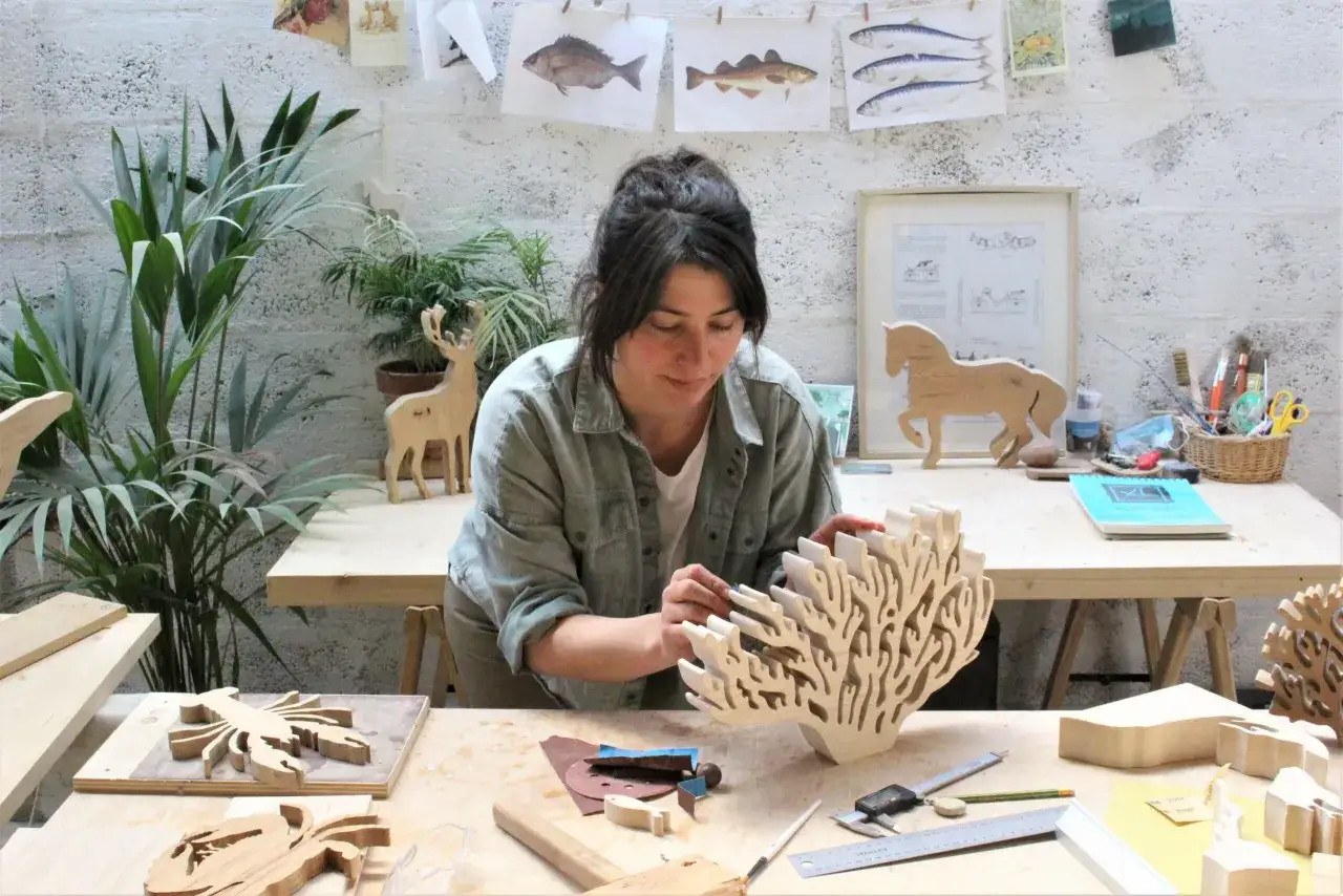 Une artiste concentrée travaille dans son atelier, entourée de ses créations en bois. Elle semble sculpter le bois avec soin, donnant vie à des formes marines et animales.