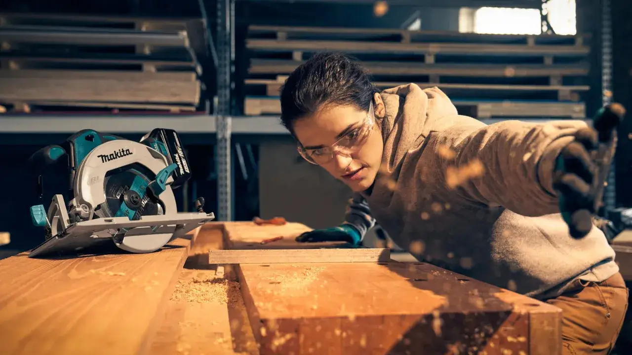 Une femme concentrée utilise une scie circulaire de marque Makita pour travailler le bois. Des copeaux volent autour d'elle.