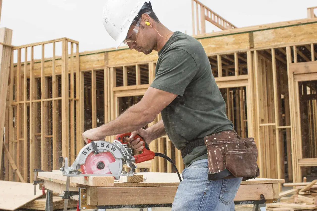Un ouvrier utilise une scie circulaire pour couper du bois sur un chantier. Il porte un casque, des lunettes et des protections auditives. Le choix de la scie circulaire est crucial pour ce travail.