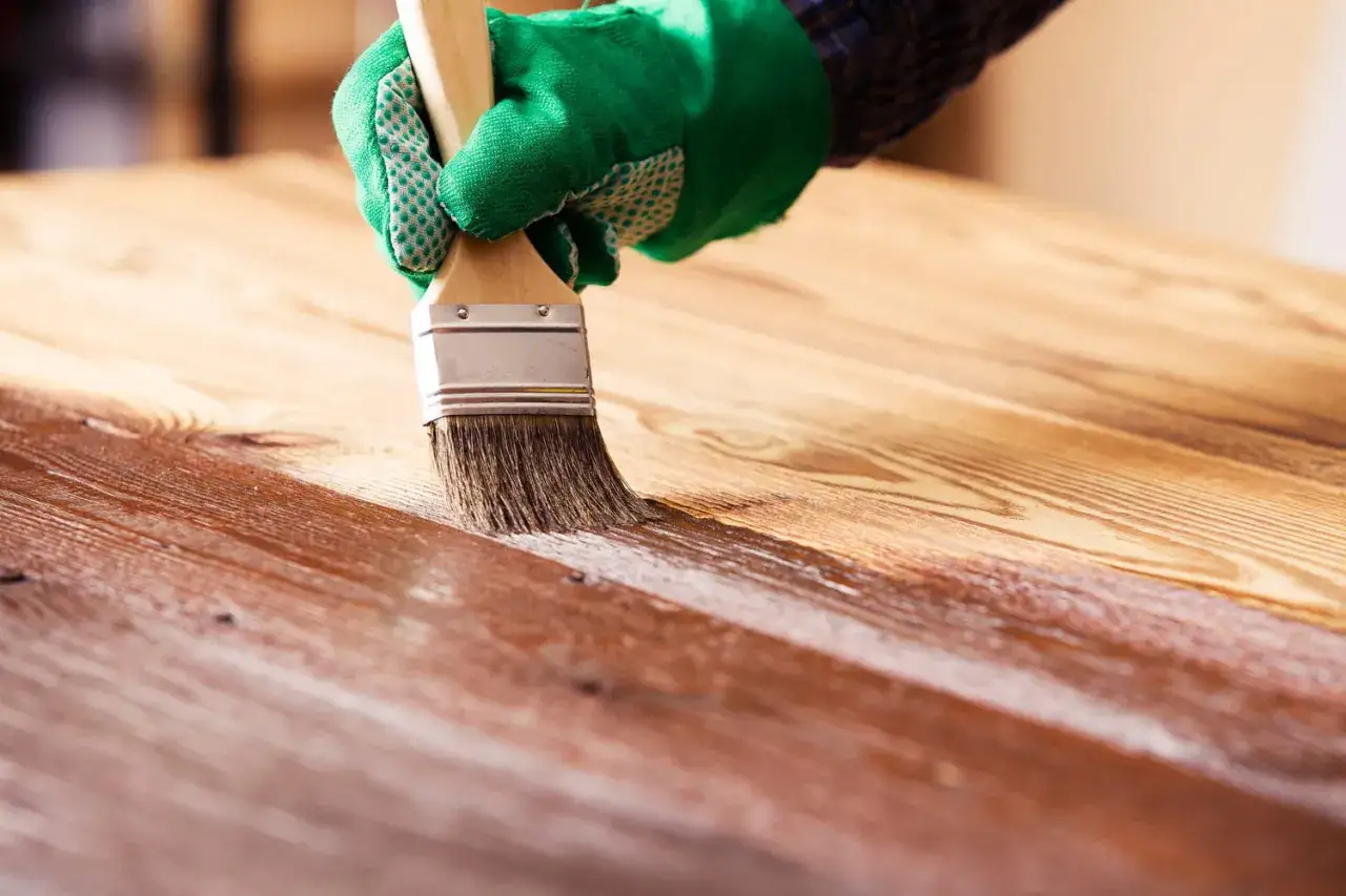 Main d'une personne portant un gant vert appliquant un vernis sur un bois vitrifié.