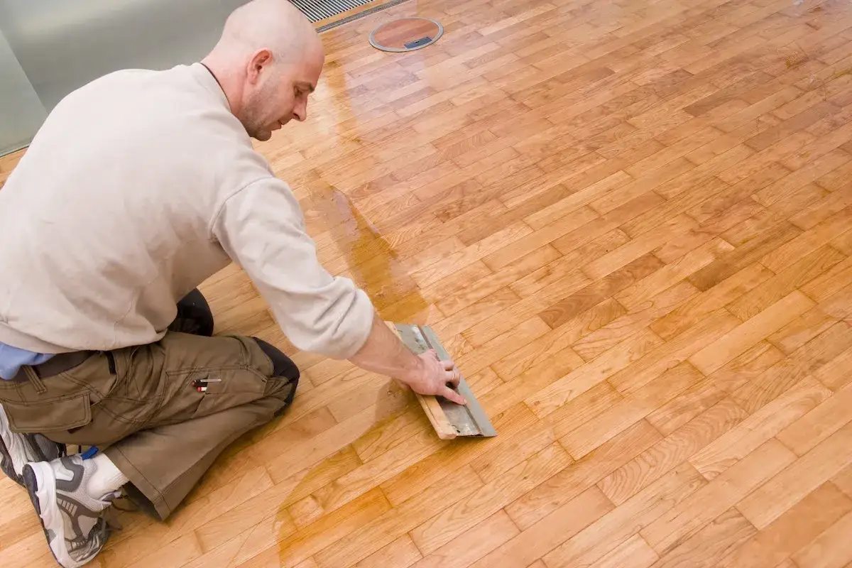 Un homme chauve, agenouill&eacute;, applique une cire sur un parquet en bois clair avec une spatule.