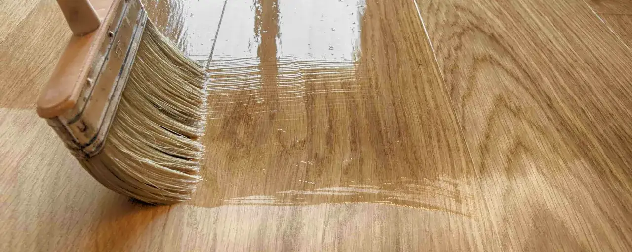 Un pinceau applique un vernis sur un parquet en bois, laissant des traces visibles. La vitrification parquet rat&eacute;e montre des irr&eacute;gularit&eacute;s et des bulles.