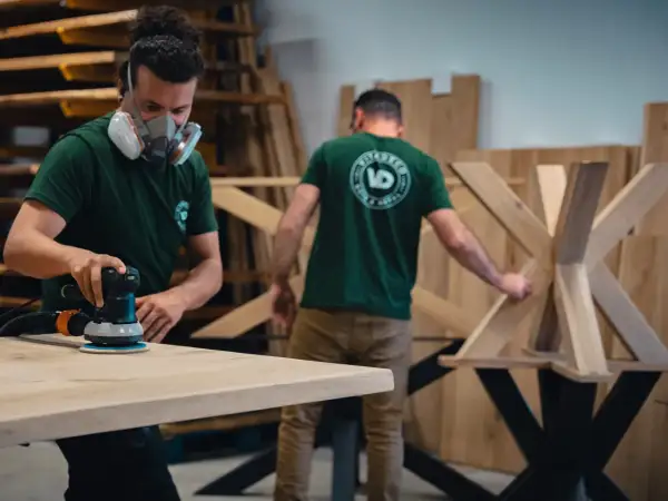 Un artisan porte un masque et utilise une ponceuse pour poncer du bois. Un autre artisan travaille sur un pied de table en bois.