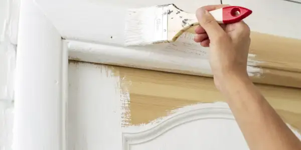 Main d'une personne peignant une porte en bois avec un pinceau. La peinture blanche recouvre partiellement le bois naturel. Quel rouleau pour peindre une porte en bois ?