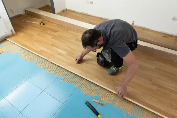 Un homme pose du parquet massif sur un sol bleu carrelé, appliquant de la colle. Il est en train de réaliser un projet de rénovation.