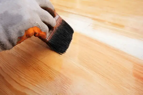 Main gauche gantée appliquant de l'huile de lin sur du bois. Attention, l'huile de lin peut présenter un danger d'auto-inflammation si mal stockée.